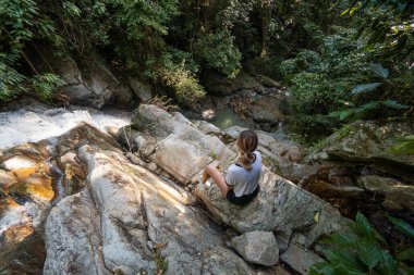 Güzel, ince, sarışın bir kadın bir kayanın üzerinde oturuyor ve ormandaki tropik şelalenin tadını çıkarıyor. Seyahat kavramı.