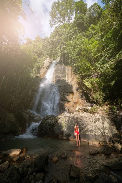 Belle jeune femme blonde mince aux cheveux bouclés, portant en maillot de bain rouge est profiter dans le lagon d'une énorme cascade tropicale dans la jungle. Concept de voyage. Femme sexy portant un maillot de bain rouge dans la jungle . — Photo