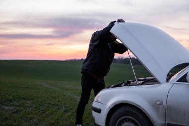Gün batımında kapüşonlu bir adam arabanın yanında duruyor. Araba arızası. Kafası karışmış adam açık kapüşonlu ve seçici yumuşak odaklı.