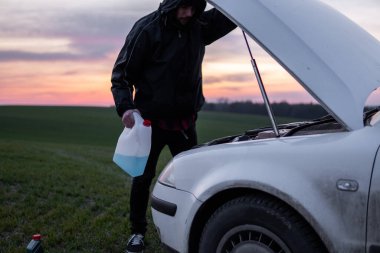 Kapüşonlu bir adam gün batımında elinde mavi sıvıyla arabanın yanında duruyor. Araba arızası. Kafası karışmış adam açık kapüşonlu ve seçici yumuşak odaklı.