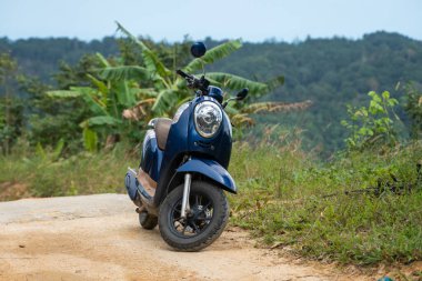 Güneşli bir günde, dağlarda manzaralı bir manzarası olan tropikal ormandaki bir tepede, toprak yolda mavi bir scooter. Macera ve seyahat konsepti.