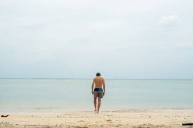 Beyaz kumsalda uzun saçlı genç bir adam tropikal denize doğru yürür. Tatil ve seyahat kavramı.