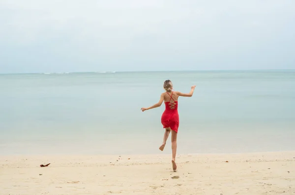 Jovem mulher em um vestido vermelho corre e salta em uma praia de areia branca em mar tropical. Jovem mulher feliz correndo na praia. Felicidade de estar em um mar ou oceano. Conceito de férias e viagens . — Fotografia de Stock