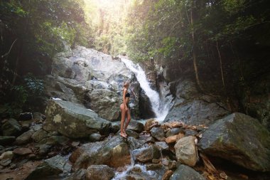 Siyah mayo giyen, kıvırcık saçlı, genç ve güzel sarışın kadın, ormandaki tropikal şelalenin oluşturduğu lagünün tadını çıkarıyor. Seyahat konsepti. Ormanda siyah mayo giyen seksi kadın..