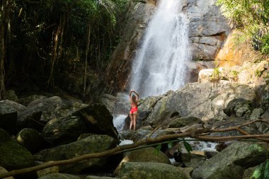 Kıvırcık saçlı, kırmızı mayo giyen genç ve güzel sarışın kadın ormandaki tropikal şelalelerin oluşturduğu lagünün tadını çıkarıyor. Seyahat konsepti. Ormanda kırmızı mayo giyen seksi kadın..