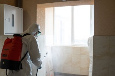 UKRAINE, KYIV - 20 Mayıs 2020: Beyaz koruyucu giysili ve maskeli adam salgının önlenmesi ve kontrol edilmesi için koronavirüs salgını sırasında iç yüzeyleri sterilize ediyor..