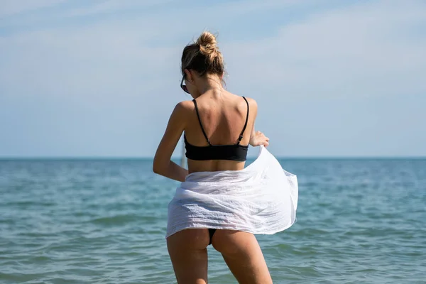 Mujer joven de pie en una playa y disfrutando del sol. Mujer joven en bikini mirando el agua de mar en la playa de resort tropical. Vista trasera. Concepto de vacaciones y viajes . —  Fotos de Stock