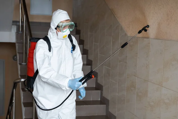 UKRAINE, KYIV - 2020年5月20日:白色の防護服とマスクを着た男が建物の内部の表面を消毒している間、感染予防と流行の制御のためのコロナウイルスの流行. — ストック写真