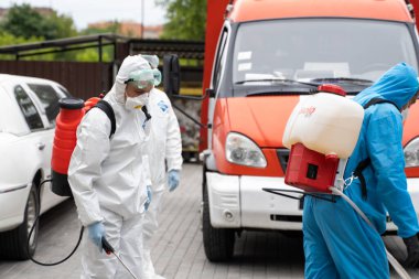 UKRAINE, KYIV - 20 Mayıs 2020: Beyaz koruyucu giysili ve maskeli bir adam bir arabanın yanında duruyor ve koronavirüs salgını sırasında iç yüzeyleri dezenfekte ediyor..