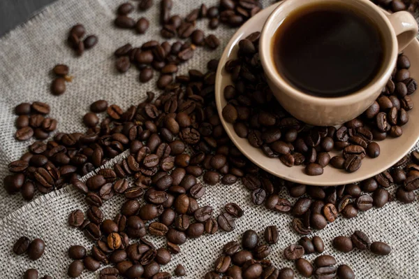 커피 잔에 볶은 커피 원두를 아마포 배경에 얹는다. 커피 가루가 뿌려진 까만 커피 잔이요. 신선 한 커피 콩. — 스톡 사진