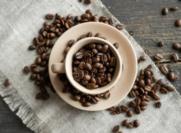 Кофейная чашка, наполненная свежей Арабикой или кофейными зёрнами робуста с разбросанными кофейными зёрнами на льняном текстильном и деревянном столе. — стоковое фото