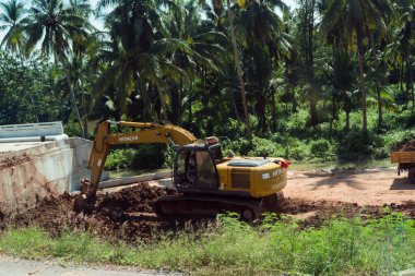 THAILAND, PHUKET - 15 Aralık 2019: Çamurda bir kazıcı duruyor. Arka planda palmiye ağaçları olan bir yol inşaatında kazıcı..