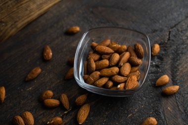 Bademler, arka plan olarak eski ahşap bir masada, tabağın etrafına saçılmış badem fındıkları olan küçük bir tabakta. Badem sağlıklı bir vejetaryen protein yiyeceğidir. Doğal çerezler.