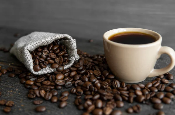 Кофе разбросан по льняной сумке и чашке черного кофе Эспрессо или Американо, стоящих на деревянном столе. Свежий арабика кофейных зерен на льняных тканях. Ароматический свежеваренный кофе. — стоковое фото