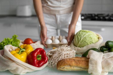 Tekrar kullanılabilir alışveriş torbalarıyla dolu yumurta tepsisini elinde tutan bir kadın evdeki bir mutfaktaki masada sebzeli. Sıfır atık ve plastiksiz konsept. Sebzeli pamuk satıcısı. Ekoloji.