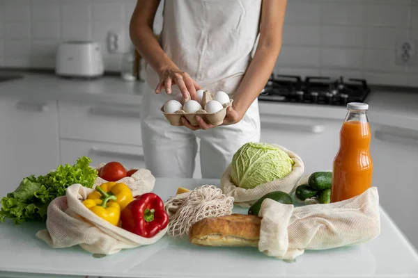 Woman holding eggs tray with a reusable shopping bags with vegetables on a table on a kitchen at home. Zero waste and plastic free concept. Mesh cotton shopper with vegetables. Ecology.