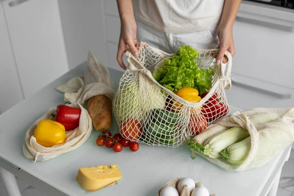Vrouw kwam terug van een markt en pakte een herbruikbare boodschappentas vol groenten uit in een keuken thuis. Geen afval en plastic vrij concept. Meisje houdt mesh katoen shopper met groenten. — Stockfoto