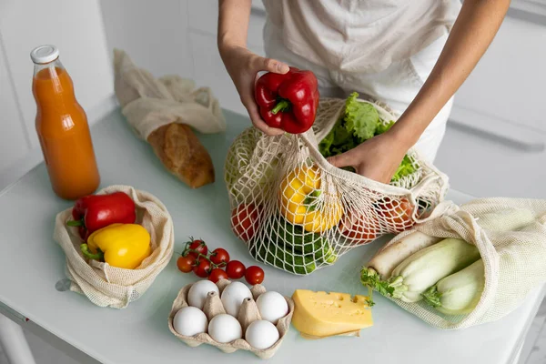 Vrouwen hand, houden een herbruikbare kruidenierszak met groenten op een keuken thuis en neemt peper uit. Geen afval en plastic vrij concept. Mesh katoenen shopper met groenten. Ecologie. — Stockfoto