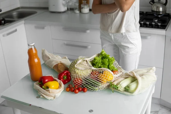 Woman came back from a market and unpacks a reusable grocery bag full of vegetables on a kitchen at home. Zero waste and plastic free concept. Girl is holding mesh cotton shopper with vegetables.