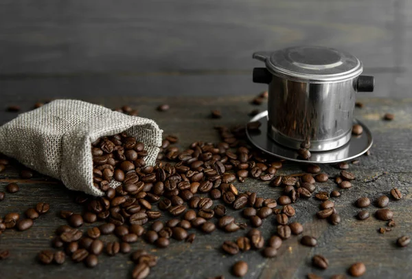 Κόκκοι καφέ διάσπαρτοι από μια λινή τσάντα σε ένα ξύλινο τραπέζι με ένα βιετναμέζικο μεταλλικό φίλτρο καφέ. Κόκκοι καφέ. — Φωτογραφία Αρχείου