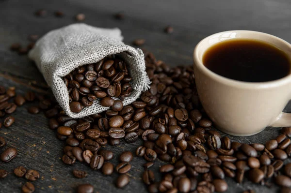 Кофе разбросан по льняной сумке и чашке черного кофе Эспрессо или Американо, стоящих на деревянном столе. Свежий арабика кофейных зерен на льняных тканях. Ароматический свежеваренный кофе. — стоковое фото
