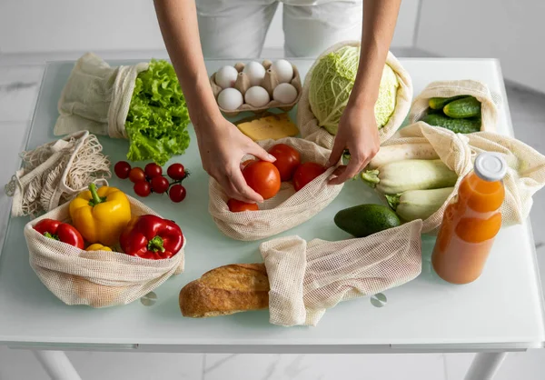 Womans hand, holding a reusable grocery bag with vegetables on a kitchen at home and takes tomato out. Zero waste and plastic free concept. Mesh cotton shopper with vegetables. Ecology. — Stock Photo, Image