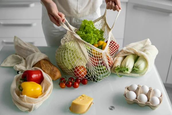 Woman came back from a market and unpacks a reusable grocery bag full of vegetables on a kitchen at home. Zero waste and plastic free concept. Girl is holding mesh cotton shopper with vegetables.
