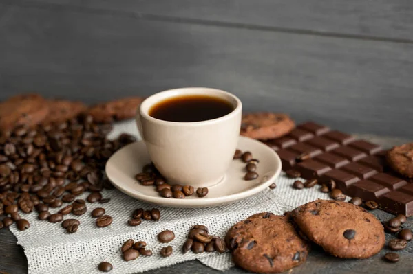 Kubek kawy z ciasteczkami i czekolady z rozproszonych ziaren kawy na pościel i drewniane tło stołu. Kubek czarnej kawy. — Zdjęcie stockowe