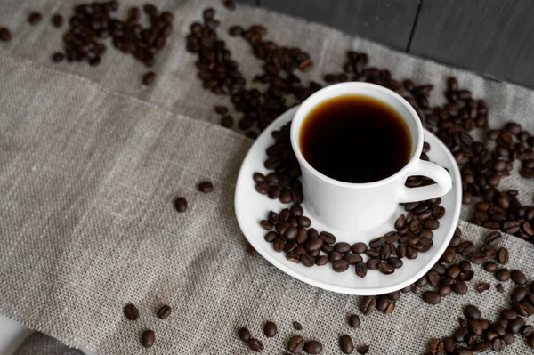 커피 잔에 볶은 커피 원두를 아마포 배경에 얹는다. 커피 가루가 뿌려진 까만 커피 잔이요. 신선 한 커피 콩. — 스톡 사진