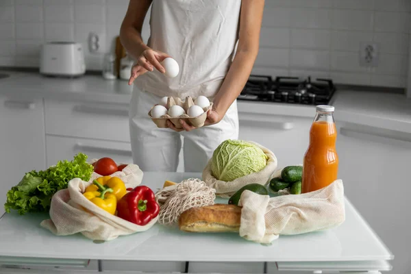 Woman holding eggs tray with a reusable shopping bags with vegetables on a table on a kitchen at home. Zero waste and plastic free concept. Mesh cotton shopper with vegetables. Ecology.