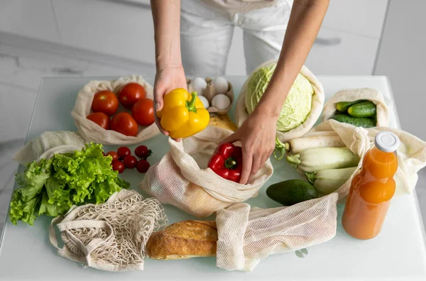 Vrouwen hand, houden een herbruikbare kruidenierszak met groenten op een keuken thuis en neemt peper uit. Geen afval en plastic vrij concept. Mesh katoenen shopper met groenten. Ecologie. — Stockfoto