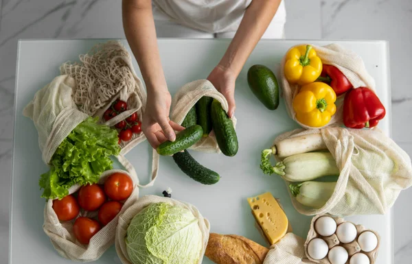 Womans hand, holding a reusable grocery bag with vegetables on a kitchen at home and takes cucumber out. Zero waste and plastic free concept. Mesh cotton shopper with vegetables. Ecology. — Stock Photo, Image