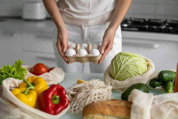 Vrouw met eierschaal met een herbruikbare boodschappentas met groenten op een tafel op een keuken thuis. Geen afval en plastic vrij concept. Mesh katoenen shopper met groenten. Ecologie. — Stockfoto
