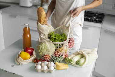 Kadın marketten döndü ve evdeki bir mutfakta sebze dolu bir poşeti açtı. Sıfır atık ve plastiksiz konsept. Kız elinde sebzeli pamuk alıcısı tutuyor..