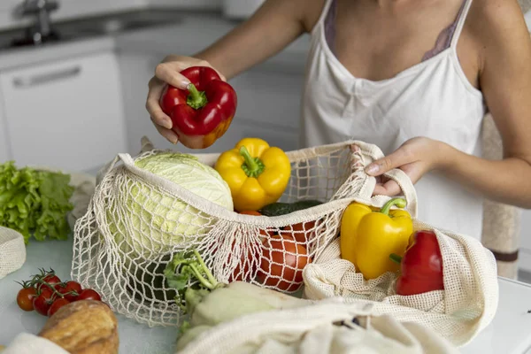 Womans hand, holding a reusable grocery bag with vegetables on a kitchen at home and takes pepper out. Zero waste and plastic free concept. Mesh cotton shopper with vegetables. Ecology. — Stock Photo, Image