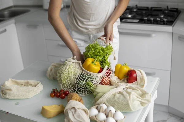 Woman came back from a market and unpacks a reusable grocery bag full of vegetables on a kitchen at home. Zero waste and plastic free concept. Girl is holding mesh cotton shopper with vegetables.