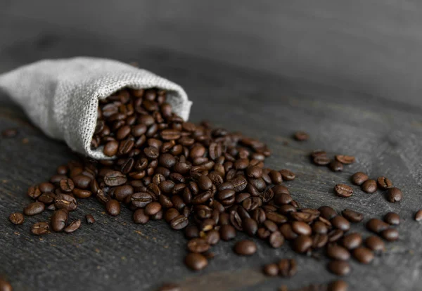 Grãos de café espalhados de uma bolsa de linho em uma mesa de madeira. Grãos de café arábica torrados frescos. — Fotografia de Stock