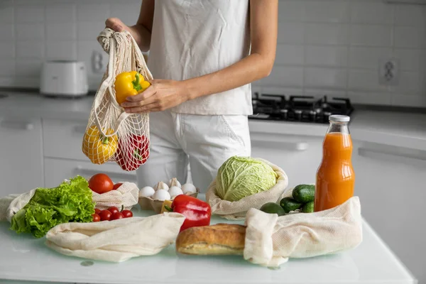 Womans hand, holding a reusable grocery bag with vegetables on a kitchen at home and takes pepper out. Zero waste and plastic free concept. Mesh cotton shopper with vegetables. Ecology. — Stock Photo, Image