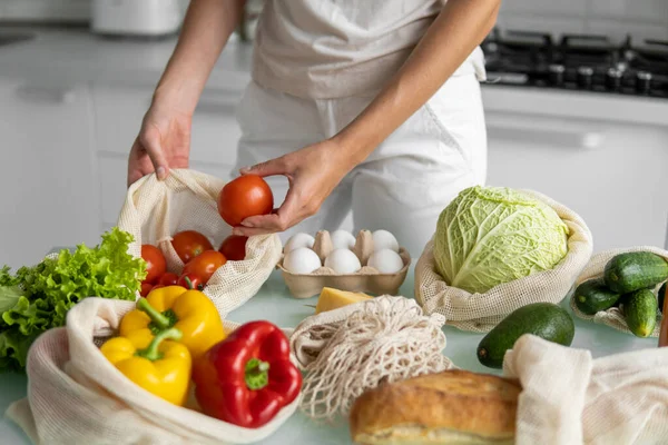 Womans hand, holding a reusable grocery bag with vegetables on a kitchen at home and takes tomato out. Zero waste and plastic free concept. Mesh cotton shopper with vegetables. Ecology. — Stock Photo, Image