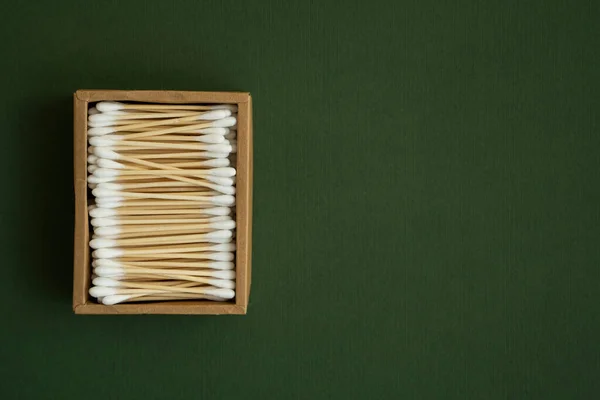 Coton-tige écologique. Une boîte en carton contenant des bourgeons de coton de bambou recyclables sur une surface verte. Concept zéro déchet. Produit écologique. Des bâtonnets de cosmétiques dans une boîte. Bâtons en bois pour nettoyer les oreilles. — Photo