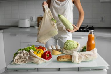 Kadın eli, evdeki mutfakta sebzeli ve tekrar kullanılabilir bir market poşeti tutuyor ve kabağı çıkarıyor. Sıfır atık ve plastiksiz konsept. Sebzeli pamuk satıcısı. Ekoloji.