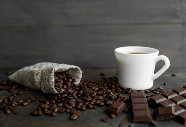 커피 원두는 나무 탁자 위에 있는 리넨 백에 뿌려져 있다. 커피 한 잔과 진한 초콜릿 바를 마시는 것입니다. 블랙 커피 머그. 초콜릿을 곁들인 아침 커피. — 스톡 사진