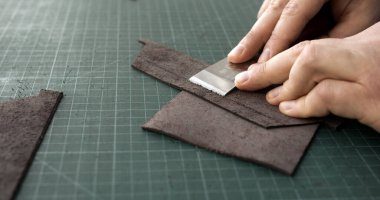 Close up of artisan hands skiving leather strip with blade tool on green cutting mat. Handmade leather goods production.