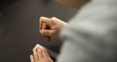 Overhead view of artisan carefully using awl and ruler to mark lines on dark leather sheet in workshop before cutting.