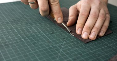 Close up of craftsman hands using beveling tool to refine leather edge on green mat. Step in handmade leather crafting for professional finish.