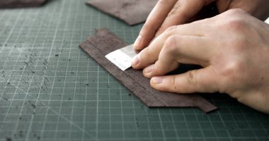 Close up of hands using sharp blade tool to thin leather strip. Traditional handmade leatherworking step performed on green cutting mat.