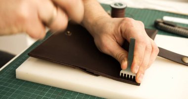 Leather artisan using mallet and pricking iron to punch stitching holes on wallet edge. Detail of handmade leatherworking process.