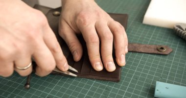 Craftsman using scissors to trim handmade wallet edges on green cutting mat. Final step of handmade leather accessory production.