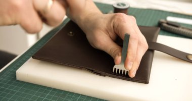 Craftsman carefully positioning pricking iron tool on leather wallet to make holes for sewing. Handmade accessory preparation.