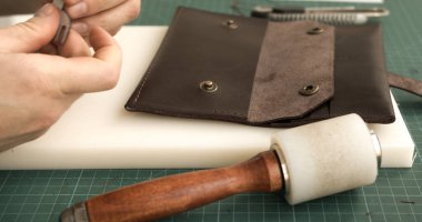 Artisan holding pricking iron and mallet while marking stitching holes on leather wallet before sewing. Handmade process step.